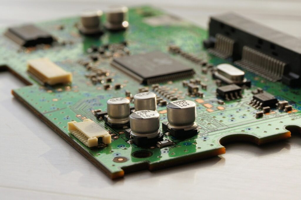 This image displays a green printed circuit board (PCB) adorned with various electronic components like capacitors and chips. Amidst the intricate circuitry, we want to let you know that "We’re Open for Business! Need PCB Layout & Design Services We've Got You Covered", ready to craft and shape PCBs to meet your specific requirements.